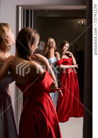 Two young beautiful girls wearing off-the-shoulder full-length sky blue and crimson red satin slit prom ball gowns. Models looking in mirror. Fitting room in dress hire service. 73741179