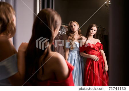 Two young beautiful girls wearing off-the-shoulder full-length sky blue and crimson red satin slit prom ball gowns. Models looking in mirror. Fitting room in dress hire service. Two young beautiful girls wearing off-the-shoulder full-length sky blue and crimson red satin slit prom ball gowns. Models looking in mirror. Fitting room in dress hire service. 73741180