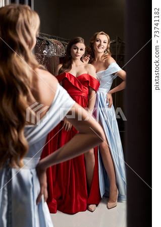 Two young beautiful girls wearing off-the-shoulder full-length sky blue and crimson red satin slit prom ball gowns. Models looking in mirror. Fitting room in dress hire service. Two young beautiful girls wearing off-the-shoulder full-length sky blue and crimson red satin slit prom ball gowns. Models looking in mirror. Fitting room in dress hire service. 73741182