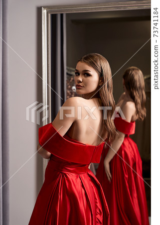 Young beautiful girl wearing an off-the-shoulder full-length crimson red satin slit prom ball gown. Model looking in mirror. Fitting room in dress hire service. Young beautiful girl wearing an off-the-shoulder full-length crimson red satin slit prom ball gown. Model looking in mirror. Fitting room in dress hire service. 73741184