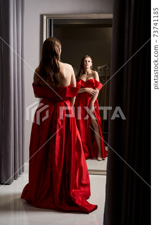 Young beautiful girl wearing an off-the-shoulder full-length crimson red satin slit prom ball gown. Model looking in mirror. Fitting room in dress hire service. Young beautiful girl wearing an off-the-shoulder full-length crimson red satin slit prom ball gown. Model looking in mirror. Fitting room in dress hire service. 73741185