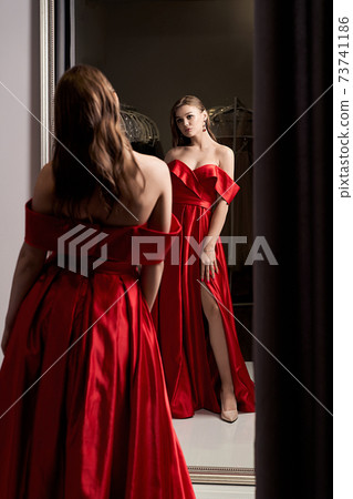 Young beautiful girl wearing an off-the-shoulder full-length crimson red satin slit prom ball gown. Model looking in mirror. Fitting room in dress hire service. Young beautiful girl wearing an off-the-shoulder full-length crimson red satin slit prom ball gown. Model looking in mirror. Fitting room in dress hire service. 73741186