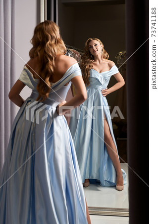 Young beautiful blonde girl wearing an off-the-shoulder full-length sky blue satin slit prom ball gown. Model looking in mirror. Fitting room in dress hire service. Young beautiful blonde girl wearing an off-the-shoulder full-length sky blue satin slit prom ball gown. Model looking in mirror. Fitting room in dress hire service. 73741188