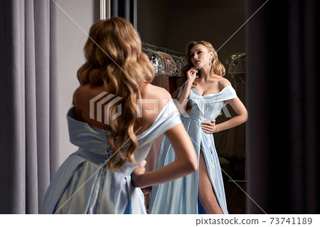 Young beautiful blonde girl wearing an off-the-shoulder full-length sky blue satin slit prom ball gown. Model looking in mirror. Fitting room in dress hire service. 73741189
