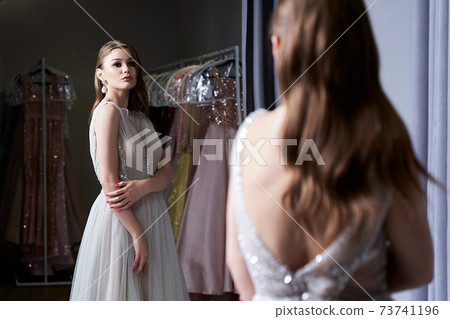 Young beautiful brunette girl wearing a full-length silver white chiffon prom ball gown decorated with sparkles and sequins. Model in front of mirror in a fitting room at dress hire service. Young beautiful brunette girl wearing a full-length silver white chiffon prom ball gown decorated with sparkles and sequins. Model in front of mirror in a fitting room at dress hire service. 73741196