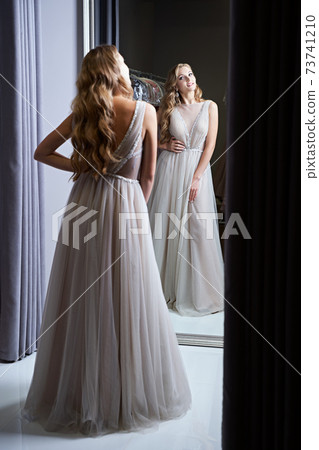 Young beautiful blonde girl wearing a full-length silver white chiffon prom ball gown decorated with sparkles and sequins. Model in front of mirror in a fitting room at dress hire service. 73741210