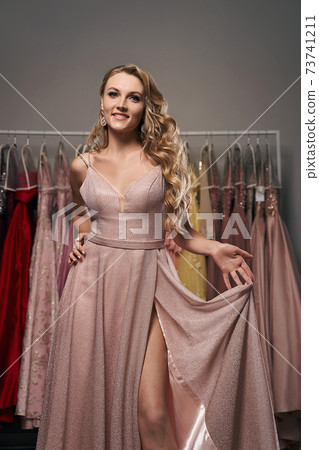 Young beautiful blonde girl wearing a full-length pale pink glitter chiffon draped prom ball gown. Model selecting an outfit for occasion in dress hire service with many options on background. Young beautiful blonde girl wearing a full-length pale pink glitter chiffon draped prom ball gown. Model selecting an outfit for occasion in dress hire service with many options on background. 73741211
