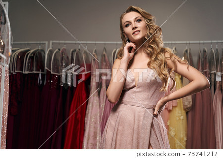 Young beautiful blonde girl wearing a full-length pale pink glitter chiffon draped prom ball gown. Model selecting an outfit for occasion in dress hire service with many options on background. Young beautiful blonde girl wearing a full-length pale pink glitter chiffon draped prom ball gown. Model selecting an outfit for occasion in dress hire service with many options on background. 73741212