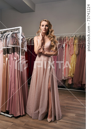 Young beautiful blonde girl wearing a full-length pale pink glitter chiffon draped prom ball gown. Model selecting an outfit for occasion in dress hire service with many options on background. Young beautiful blonde girl wearing a full-length pale pink glitter chiffon draped prom ball gown. Model selecting an outfit for occasion in dress hire service with many options on background. 73741224