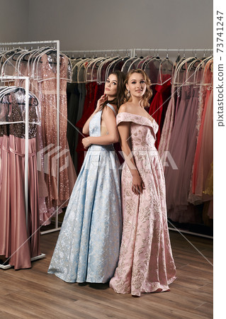 Two young beautiful girls wearing full-length draped sky blue and pale pink satin slit prom ball gowns decorated with embroindered patterns. Dress hire service. 73741247