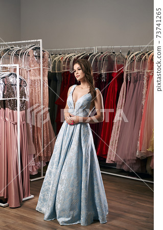 Young beautiful brunette girl wearing a full-length draped sky blue satin slit prom ball gown decorated with embroindered roses pattern. Dress hire service with many dresses on background. 73741265
