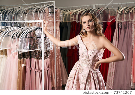 Young beautiful blonde girl wearing a full-length draped one shoulder pale pink satin slit prom ball gown decorated with embroindered pattern. Dress rental service with many dresses in on background. Young beautiful blonde girl wearing a full-length draped one shoulder pale pink satin slit prom ball gown decorated with embroindered pattern. Dress rental service with many dresses in on background. 73741267