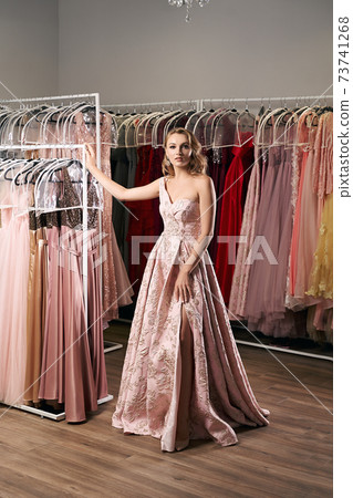 Young beautiful blonde girl wearing a full-length draped one shoulder pale pink satin slit prom ball gown decorated with embroindered pattern. Dress rental service with many dresses in on background. Young beautiful blonde girl wearing a full-length draped one shoulder pale pink satin slit prom ball gown decorated with embroindered pattern. Dress rental service with many dresses in on background. 73741268