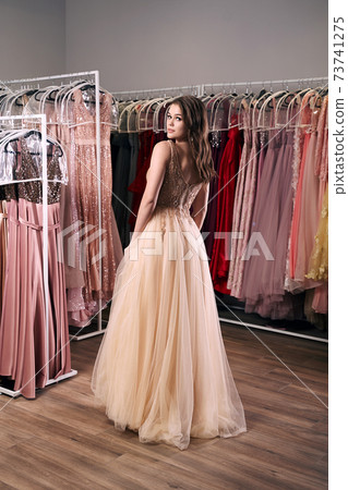 Young beautiful brunette girl wearing a full-length beige yellow champaign chiffon slit prom ball gown decorated with golden sparkles and sequins. Dress hire service with many dresses on background. Young beautiful brunette girl wearing a full-length beige yellow champaign chiffon slit prom ball gown decorated with golden sparkles and sequins. Dress hire service with many dresses on background. 73741275