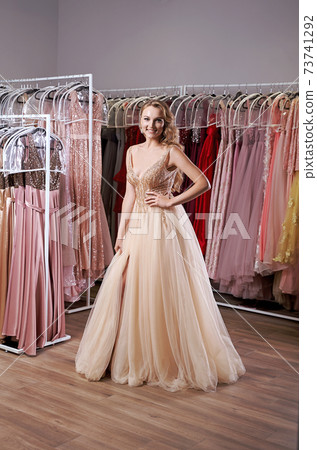 Young beautiful blonde girl wearing a full-length beige yellow champaign chiffon slit prom ball gown decorated with golden sparkles and sequins. Fashion model in dress hire service with many dresses. 73741292