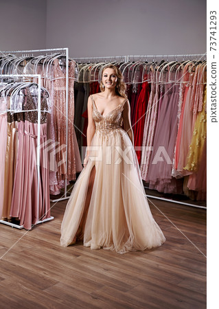 Young beautiful blonde girl wearing a full-length beige yellow champaign chiffon slit prom ball gown decorated with golden sparkles and sequins. Fashion model in dress hire service with many dresses. Young beautiful blonde girl wearing a full-length beige yellow champaign chiffon slit prom ball gown decorated with golden sparkles and sequins. Fashion model in dress hire service with many dresses. 73741293