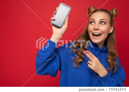 Attractive smiling young blonde woman wearing stylish blue hoodie isolated on red background with Attractive smiling young blonde woman wearing stylish blue hoodie isolated on red background with 73741473