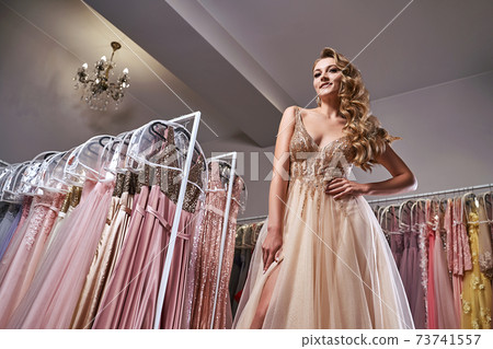 Beautiful blonde girl wearing a full-length yellow champaign chiffon slit prom ball gown decorated with golden sequins. Model in dress hire service with many dresses in different color and style. 73741557