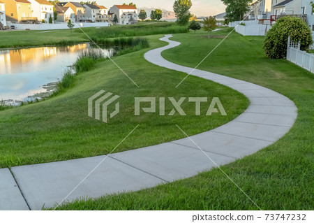 Winding footpath along a shiny pond in front of homes viewed on a sunny day 73747232