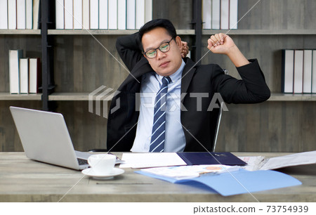 A businessman in a suit stretches lazily while looking at a computer screen. To alleviate pain from prolonged computer use 73754939