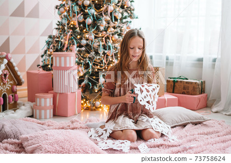 Adorable little girl sitting near the tree and making paper snow-flakes. Room decorated. Adorable little girl sitting near the tree and making paper snow-flakes. Room decorated. 73758624