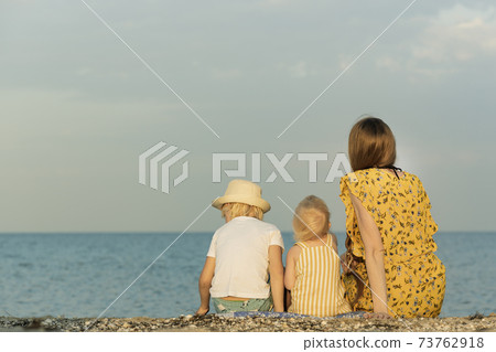 Mother with two children sitting on the beach and looking out to sea. Back view. Summer vacation with children 73762918