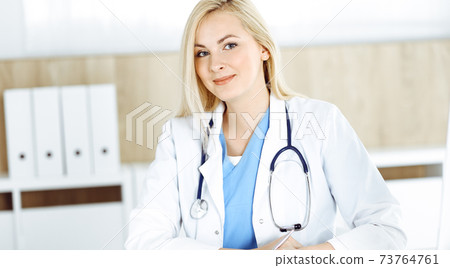 Woman-doctor at work while sitting at the desk in hospital or clinic. Blonde cheerful physician filling up medication history record form Woman-doctor at work while sitting at the desk in hospital or clinic. Blonde cheerful physician filling up medication history record form 73764761