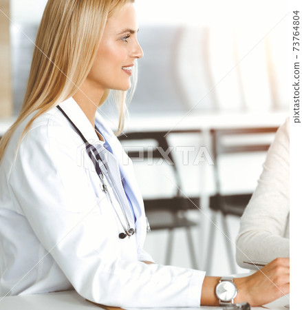 Woman-doctor at work in sunny hospital is happy to consult female patient. Blonde physician checks medical history record and exam results while using clipboard Woman-doctor at work in sunny hospital is happy to consult female patient. Blonde physician checks medical history record and exam results while using clipboard 73764804