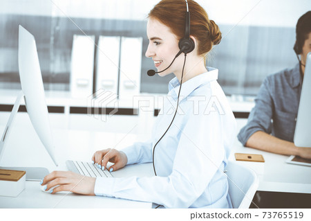 Young woman using headset and computer while talking with customers online. Group of Call center operators at work. Business concept 73765519