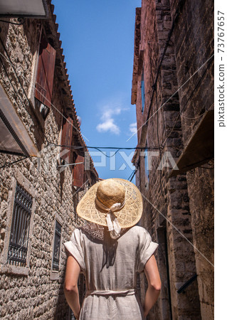 Rear view of beautiful blonde young female traveler wearing straw sun hat sightseeing and enjoying summer vacation in an old traditional costal town at Adriatic cost, Croatia 73767657