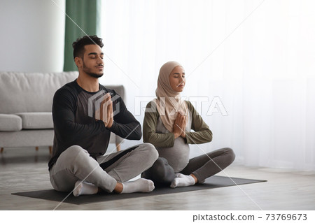 Pregnant Muslim Woman In Hijab Meditating Together With Her Husband At Home Pregnant Muslim Woman In Hijab Meditating Together With Her Husband At Home 73769673