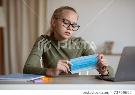 School girl holding face mask, sitting in front of laptop 73769693