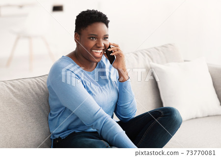 Black lady calling her friend, sitting on couch Black lady calling her friend, sitting on couch 73770081