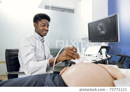 Close-up of African man doctor sonographer scanning young pregnant woman's belly with ultrasonic transducer doing obstetric ultrasonography 73771121