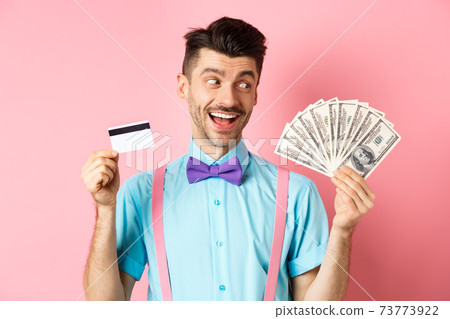 Image of smiling young man in bow-tie holding plastic credit card and looking happy at money, choosing cash, standing over pink background 73773922