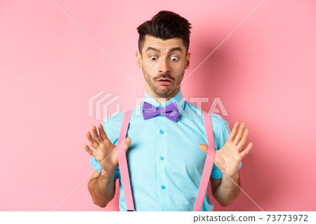Funny guy with moustache and bow-tie, adjusting his suspenders and looking down with confused and surprised face, standing over pink background 73773972
