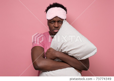 African young man looks sleepy and angry, holding pillow in hands. African young man looks sleepy and angry, holding pillow in hands. 73774349