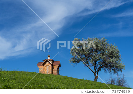 Small old wooden chapel on summer green grassy hill top, lonely willow tree and blue sky with cloud. 73774929