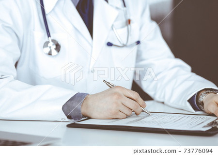 Unknown male doctor sitting and working with clipboard in clinic at his working place, close-up. Young physician at work. Perfect medical service, medicine concept Unknown male doctor sitting and working with clipboard in clinic at his working place, close-up. Young physician at work. Perfect medical service, medicine concept 73776094