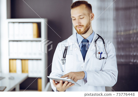 Friendly red-bearded doctor using tablet computer in clinic at his working place. Perfect medical service in hospital and medicine concept Friendly red-bearded doctor using tablet computer in clinic at his working place. Perfect medical service in hospital and medicine concept 73776448