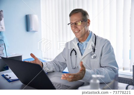 Happy old male doctor consulting patient online by video call on laptop. 73778221