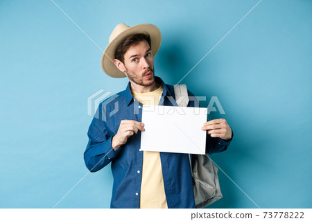 Excited tourist in straw hat, hitchhiking, showing blank piece of paper and looking amused, standing on blue background 73778222