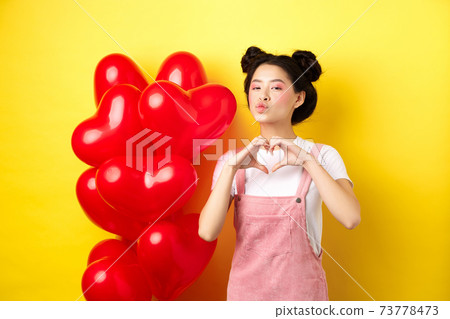 Beautiful asian woman showing heart, I love you gesture and kissing lips, standing near romantic red balloons. Concept of Valentines day 73778473