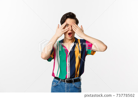 Image of handsome young gay man open eyes to see surprise, peeking through fingers with amazed emotion, standing over white background 73778502