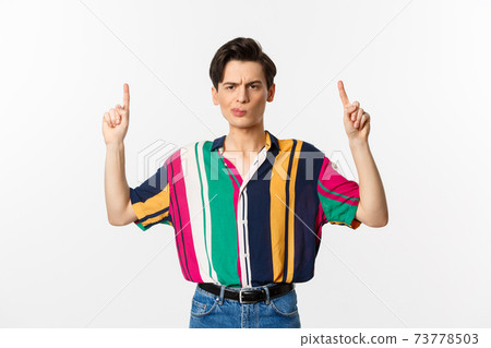 Skeptical and displeased young man pointing fingers up, grimacing disappointed, standing against white background 73778503