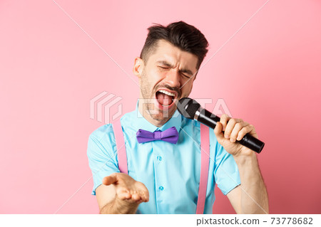 Party and festive events concept. Passionate singer holding microphone and pointing hand at you, singing about love, standing on pink background Party and festive events concept. Passionate singer holding microphone and pointing hand at you, singing about love, standing on pink background 73778682