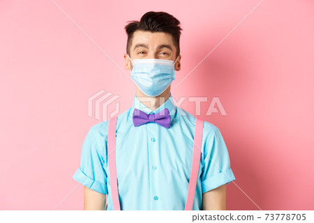Covid-19, pandemic and health concept. Portrait of smiling man in medical mask feeling happy, standing in festive bow-tie and suspenders, pink background 73778705