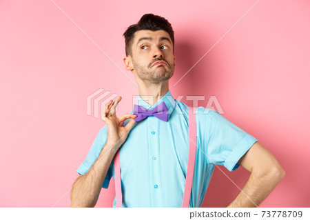 Image of funny man raising chin up and adjusting bow-tie, standing confident and sassy on pink background, looking left 73778709