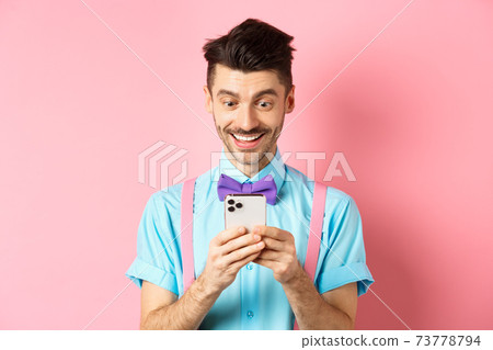 Online shopping. Happy guy looking at smartphone screen, reading message and smiling, standing on pink background 73778794