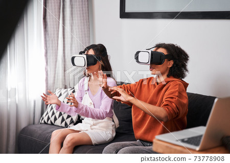Besties sitting on cozy sofa and using virtual reality goggles 73778908